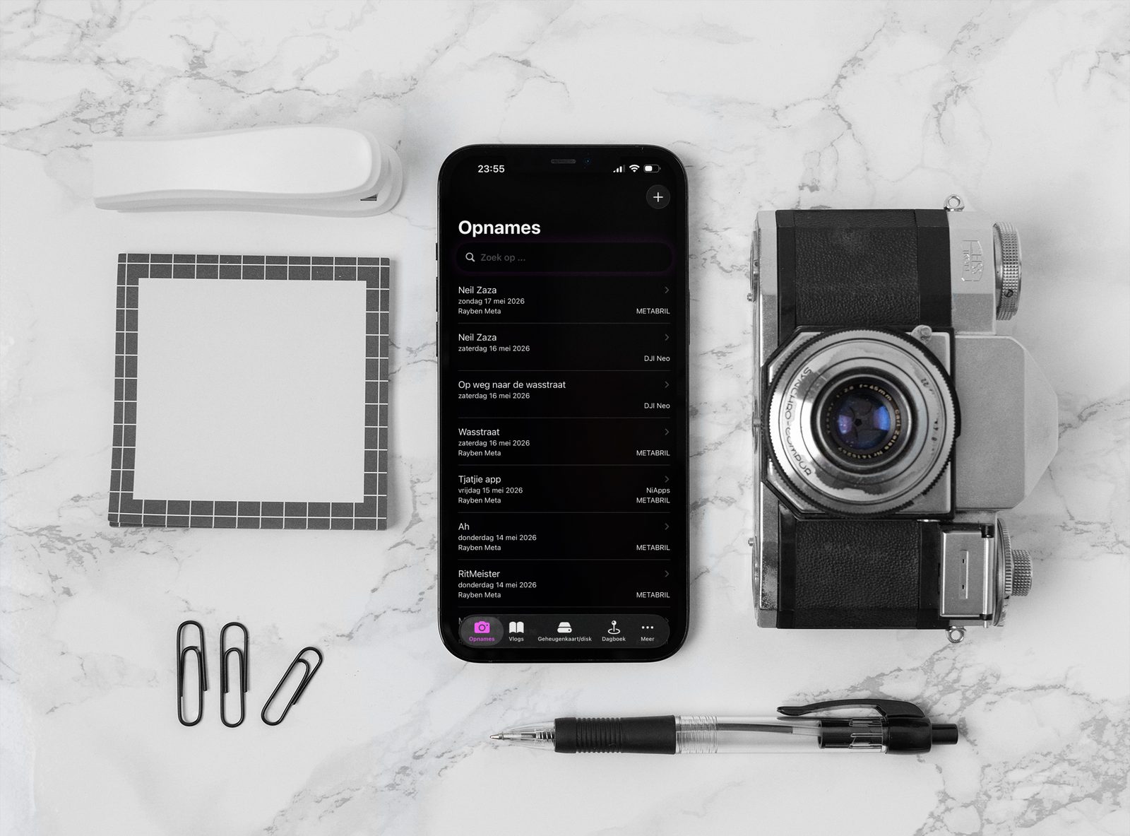 FilmSticks app on iPhone laid flat on white marble next to a vintage film camera and notepad, showing the recordings list view
