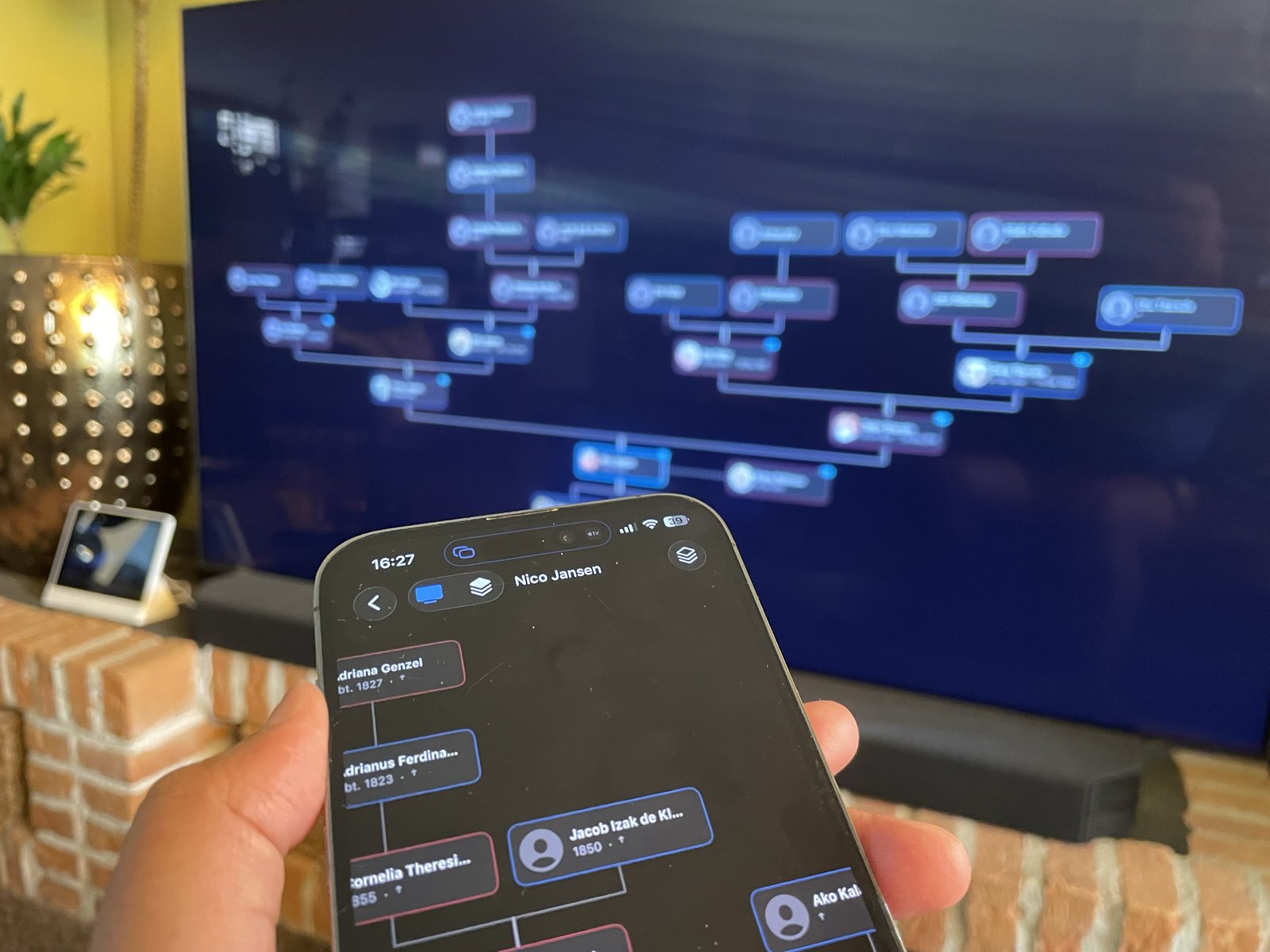 A hand holding an iPhone with the Gramps2 chart, navigating the same family tree shown large on an Apple TV in the living room