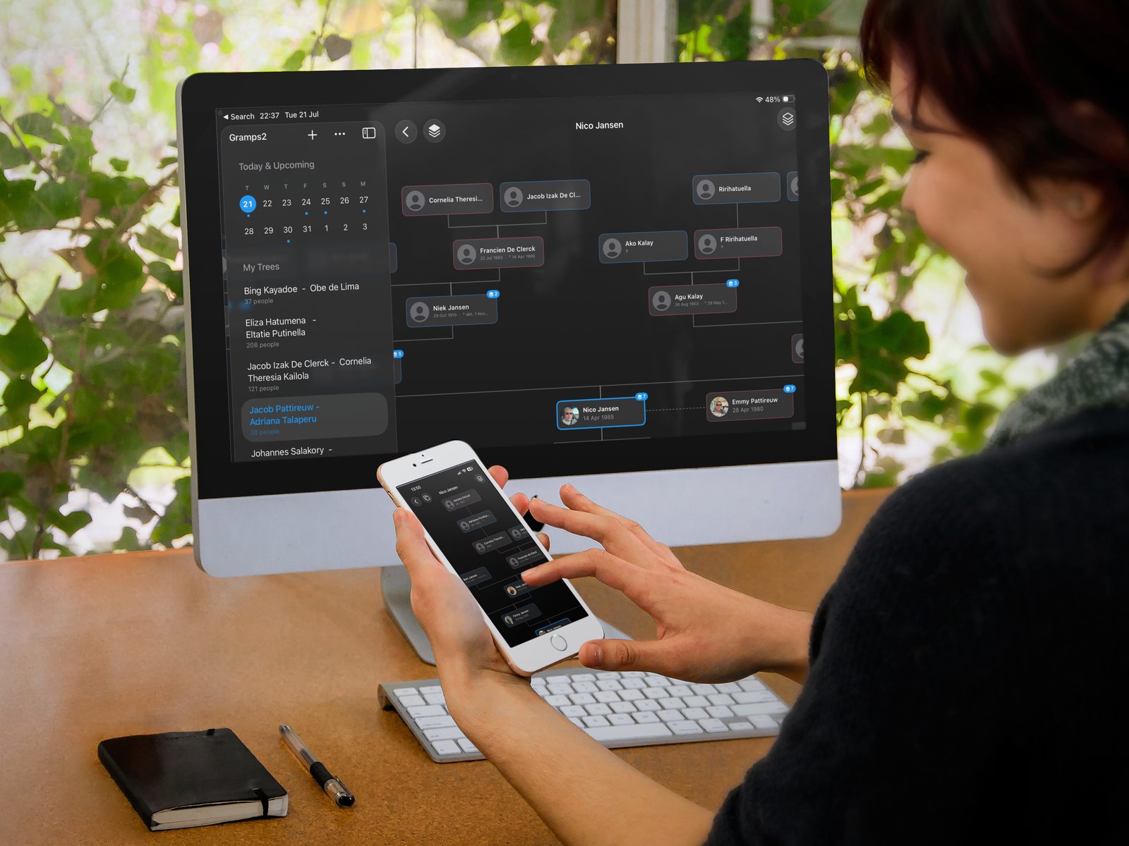 A woman exploring a Gramps2 family tree on her iPhone at a desk, with the same tree open large on a desktop screen