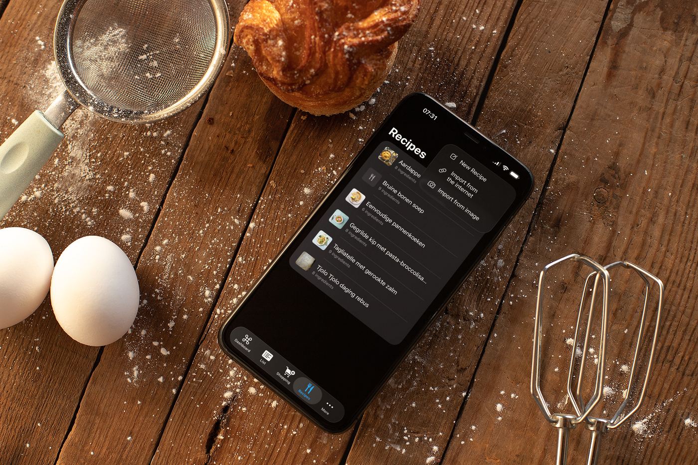 Tjatjie app on iPhone surrounded by baking supplies on a wooden surface, showing a list of recipes including Boterkoek and banana cake