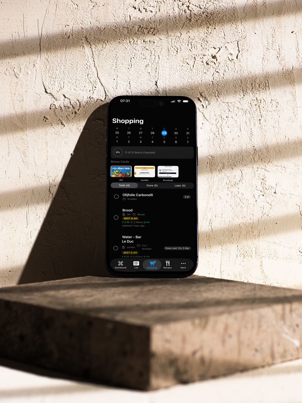 Tjatjie app on iPhone propped against a textured concrete wall, showing a shopping list with bonus cards and items including olive oil and bread